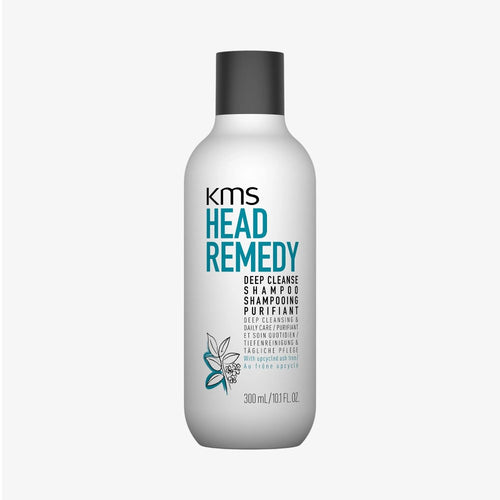 Foto di prodotto del flacone bianco di shampoo KMS Head Remedy Deep Cleanse Purifiant con tappo grigio scuro scritte principali blu e nere illustrazione di foglie stilizzate turchesi e grigie e l'indicazione di frassino riciclato su sfondo chiaro